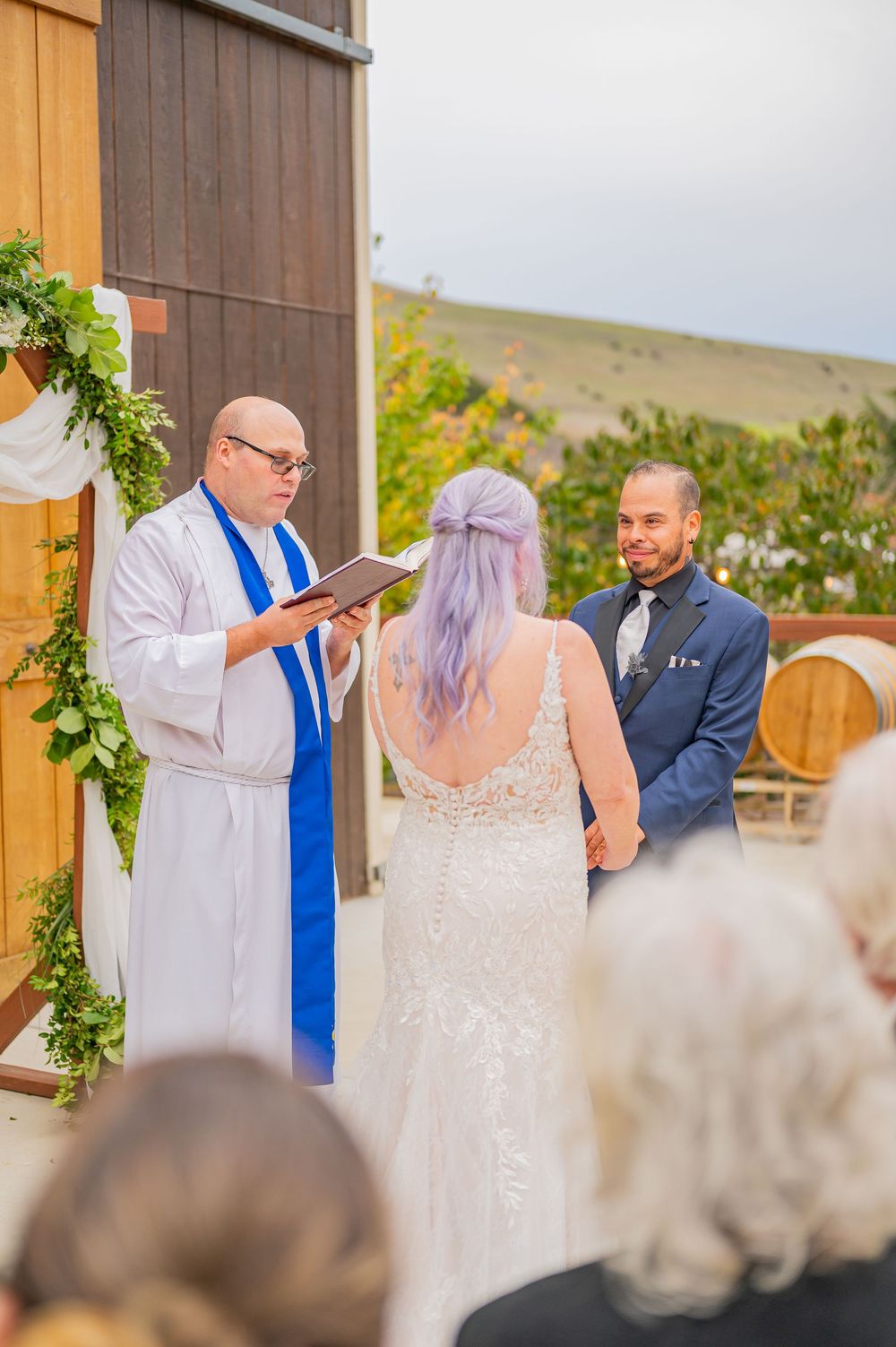 Bride with purple hair and groom holding hands at outdoor barn wedding ceremony with officiant reading vows.