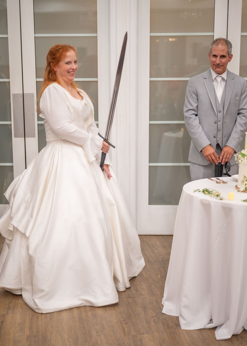 Bride in white gown holds a sword at wedding cake cutting ceremony while groom in gray suit watches.