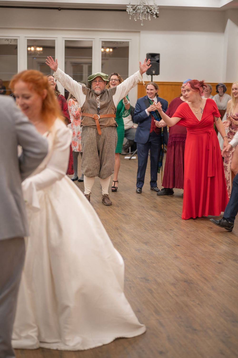 Wedding reception dancing with bride in white gown, guests in costumes and formal wear clapping on a wooden dance floor.