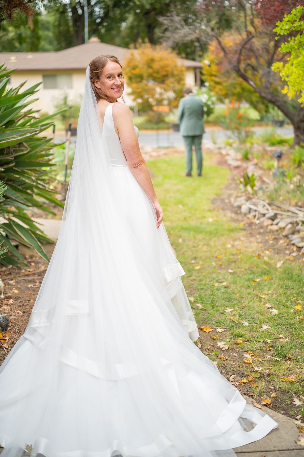 Bride in white gown and veil smiles over shoulder in garden, groom stands in background amid autumn trees.