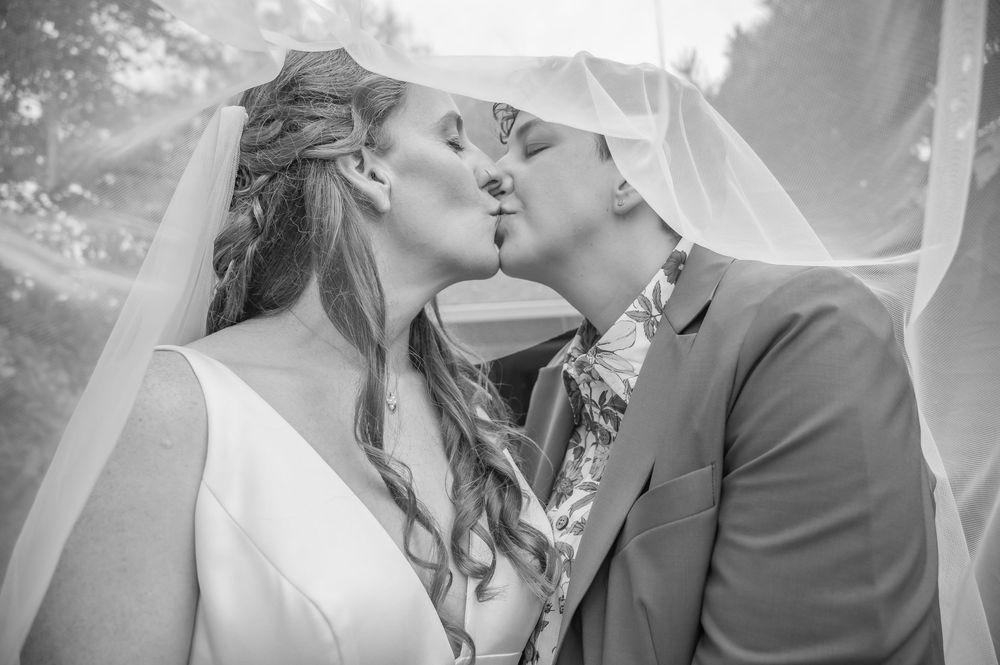 Black and white photo of a bride and groom sharing a romantic kiss under a flowing veil on their wedding day.