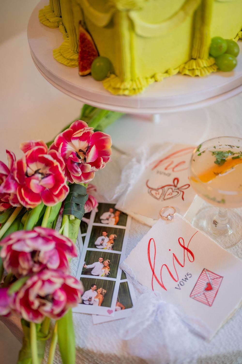 Wedding flat lay with pink tulips, vow booklets, Polaroids, cocktail, and a lime green ruffle cake on a white stand.