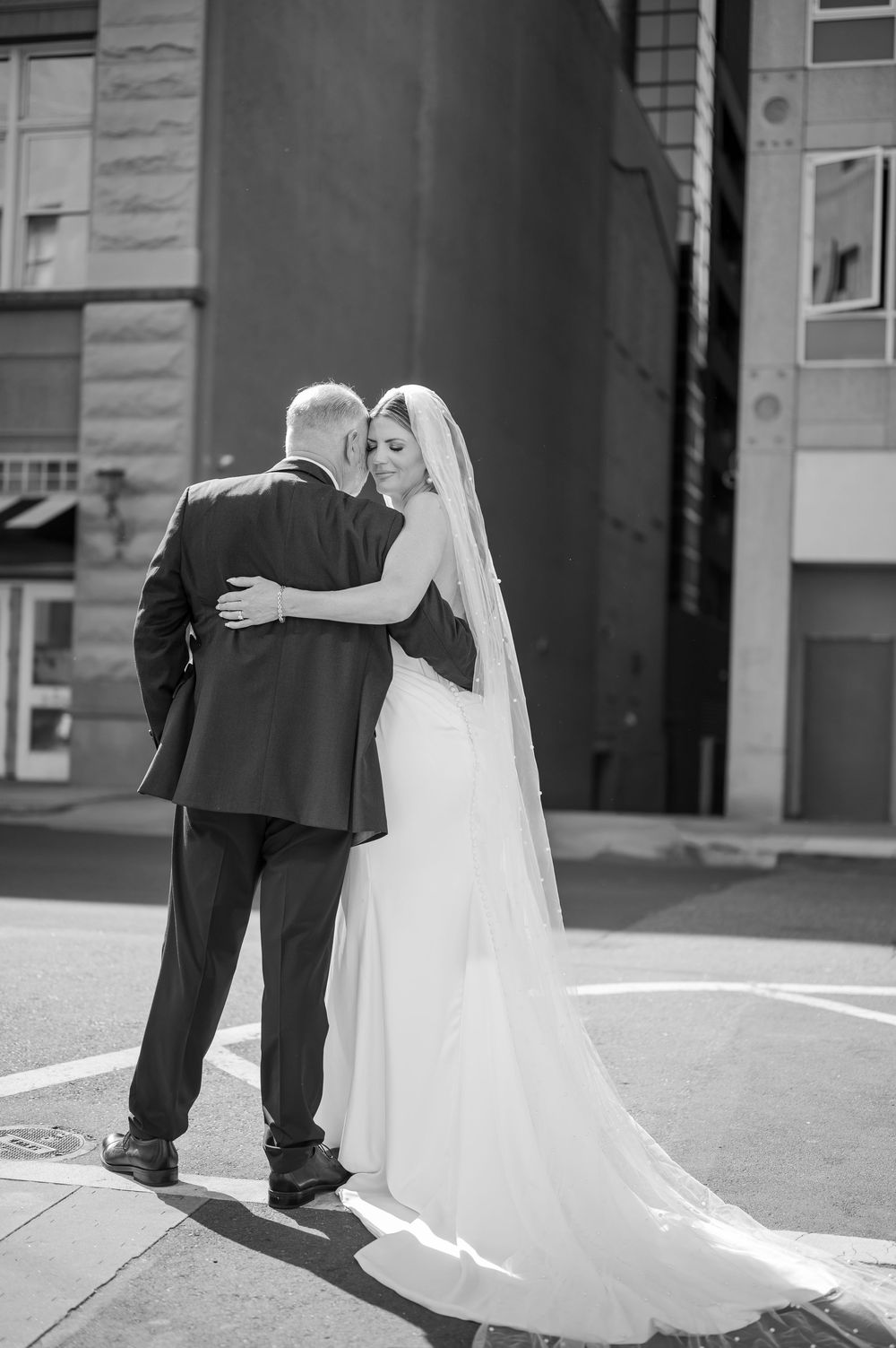 Black and white photo of a bride and groom embracing on a city street, her veil trailing behind in a romantic wedding portrait.