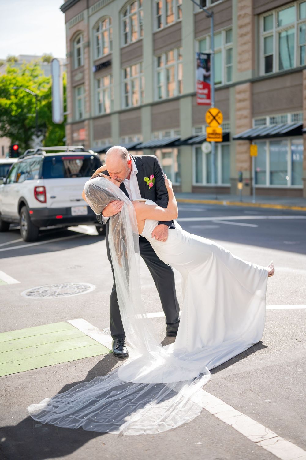 Groom dips bride in a romantic kiss on a sunny city street, her long white veil trailing on the ground.