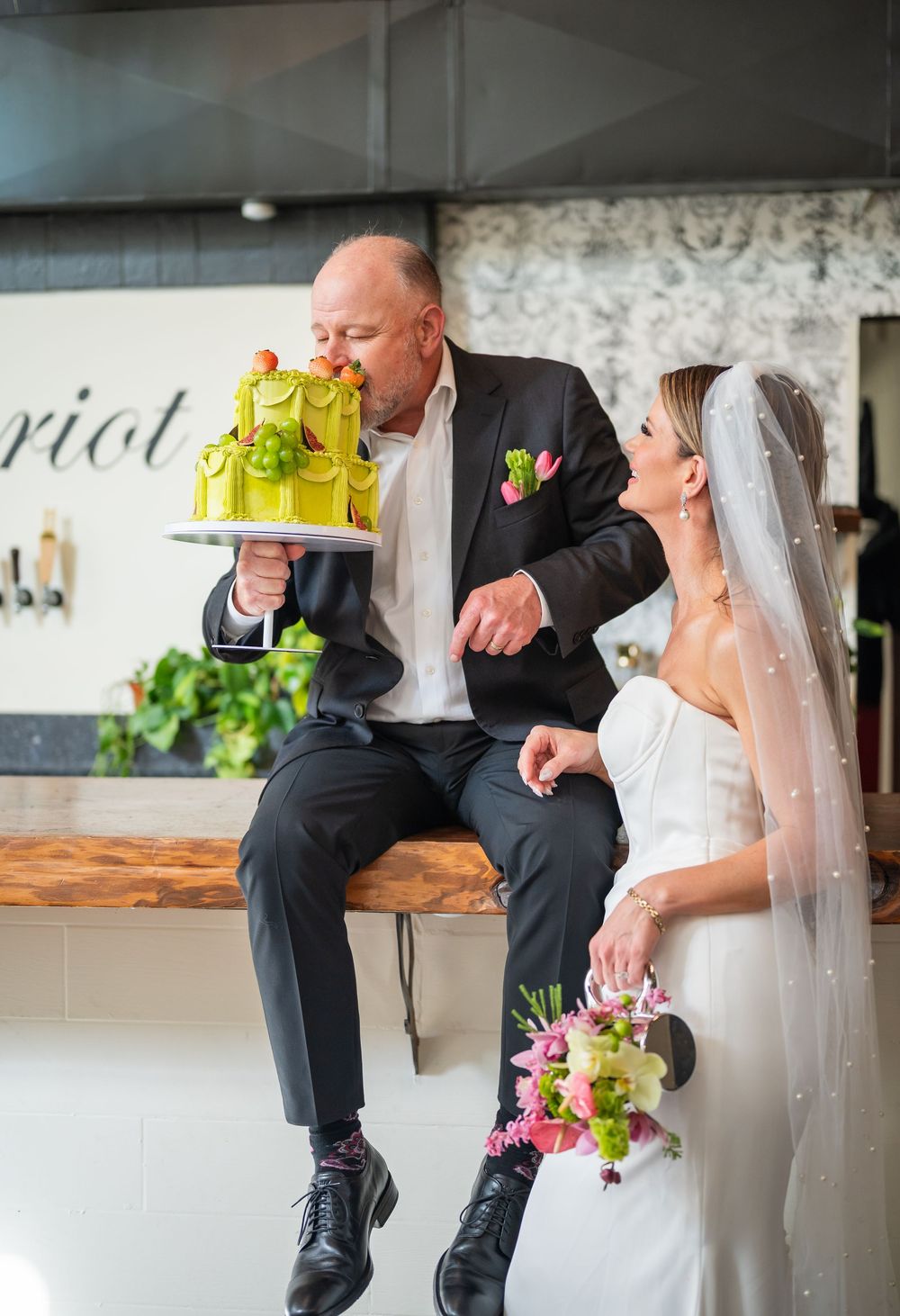 Groom kisses green wedding cake while bride in white gown and veil holds floral bouquet at wedding reception.