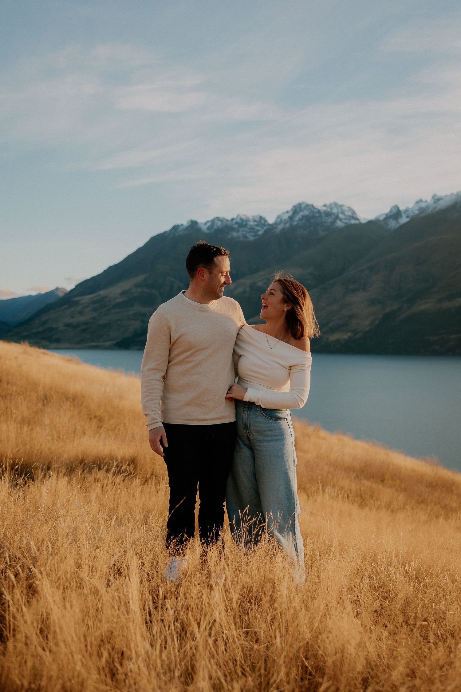 Couple embracing in golden grass field with snow-capped mountains and lake backdrop in New Zealand.