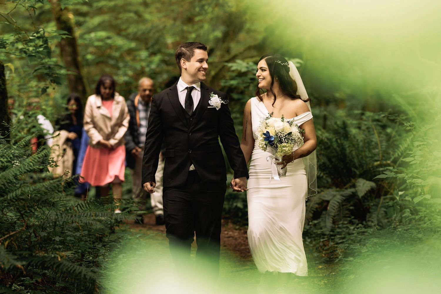 Bride and groom walk hand in hand through a lush green forest during their outdoor wedding ceremony.