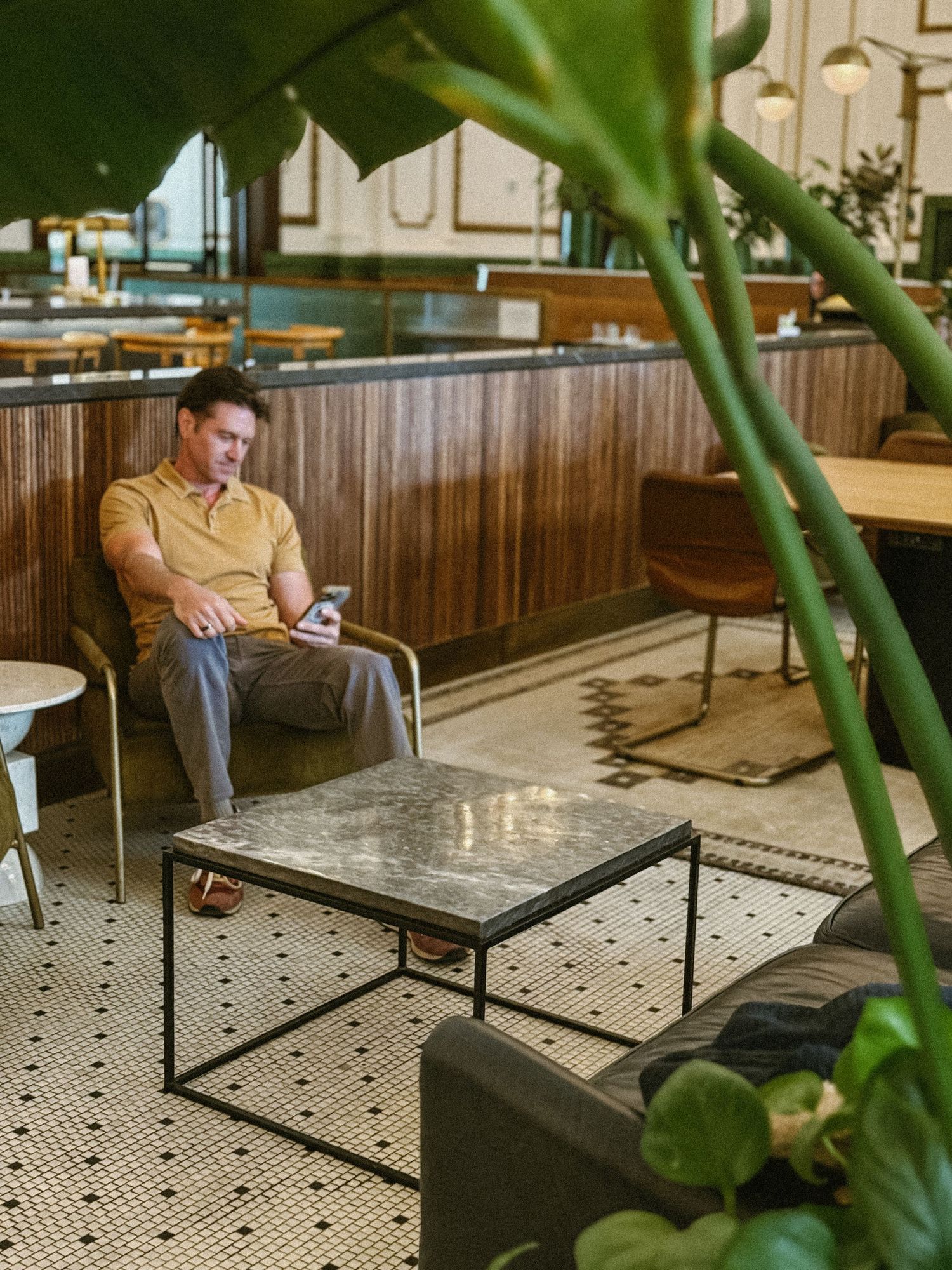 Man in yellow polo sitting in modern lounge chair using smartphone surrounded by tropical plants.