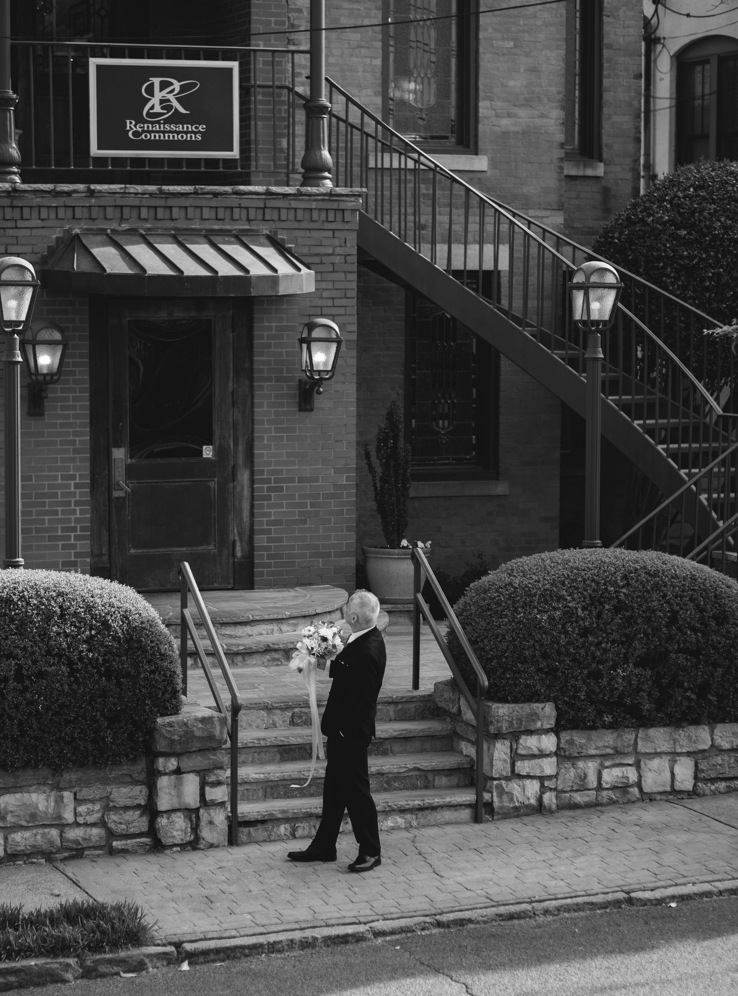 Man in tuxedo carrying bouquet walks past brick building with iron staircase in black and white photo.