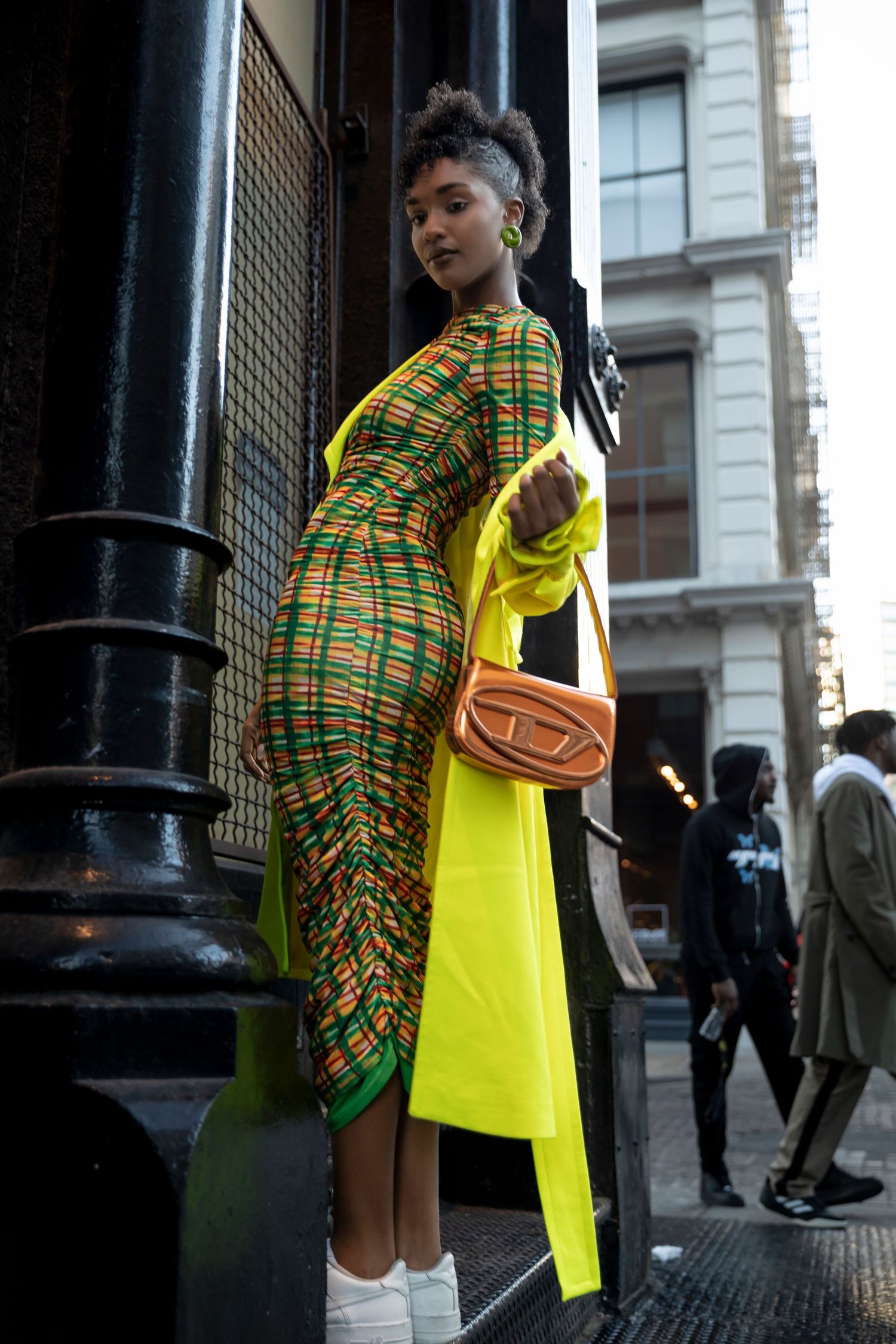 Woman in colorful plaid midi dress with neon yellow coat and orange Diesel bag posing on urban street.