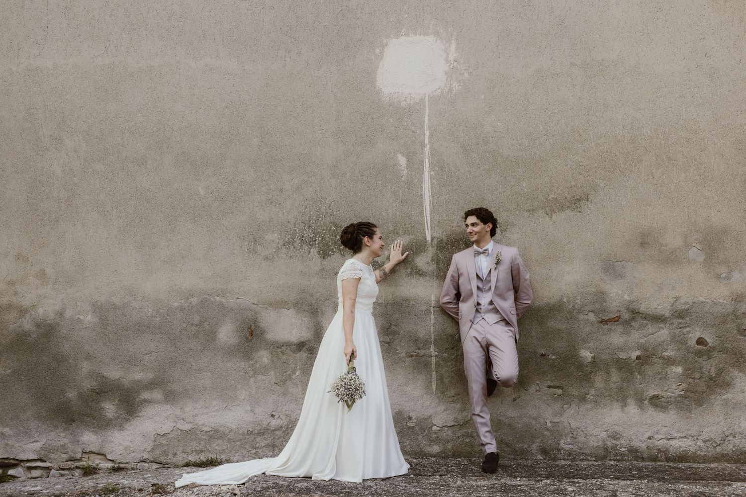 Bride and groom posing by a rustic wall with a painted balloon, sharing a romantic moment on their wedding day.