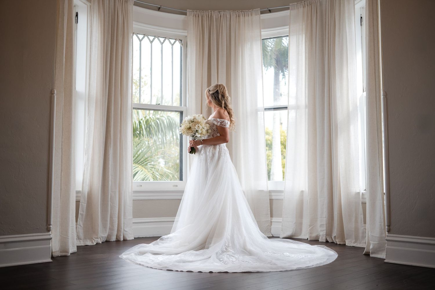 Bride in white wedding gown holding bouquet stands before elegant bay windows with sheer curtains in a beautiful venue.