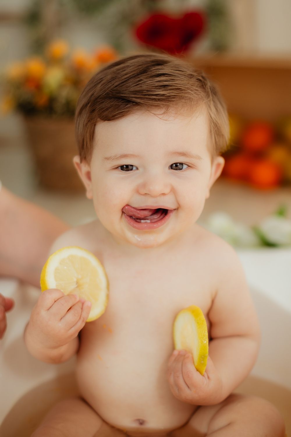 Smiling baby holding two lemon slices with tongue out, sitting in a bath surrounded by colorful fruits and flowers.