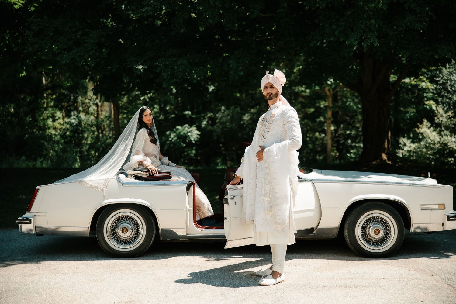 Sikh bride and groom in white traditional attire posing with a vintage cream convertible car outdoors.