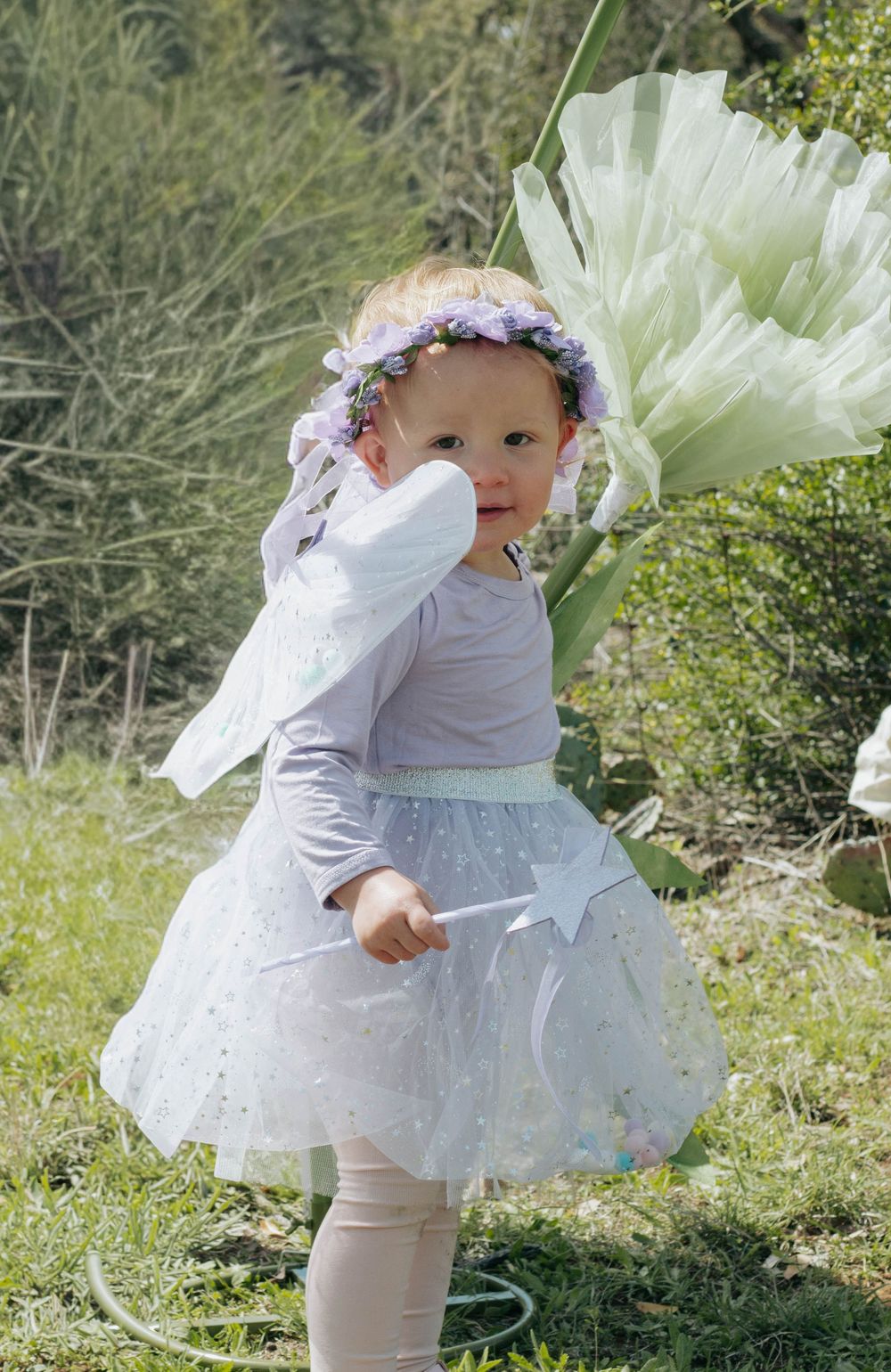 Toddler girl in fairy costume with tutu skirt, wings, floral crown, and wand standing outdoors in garden.