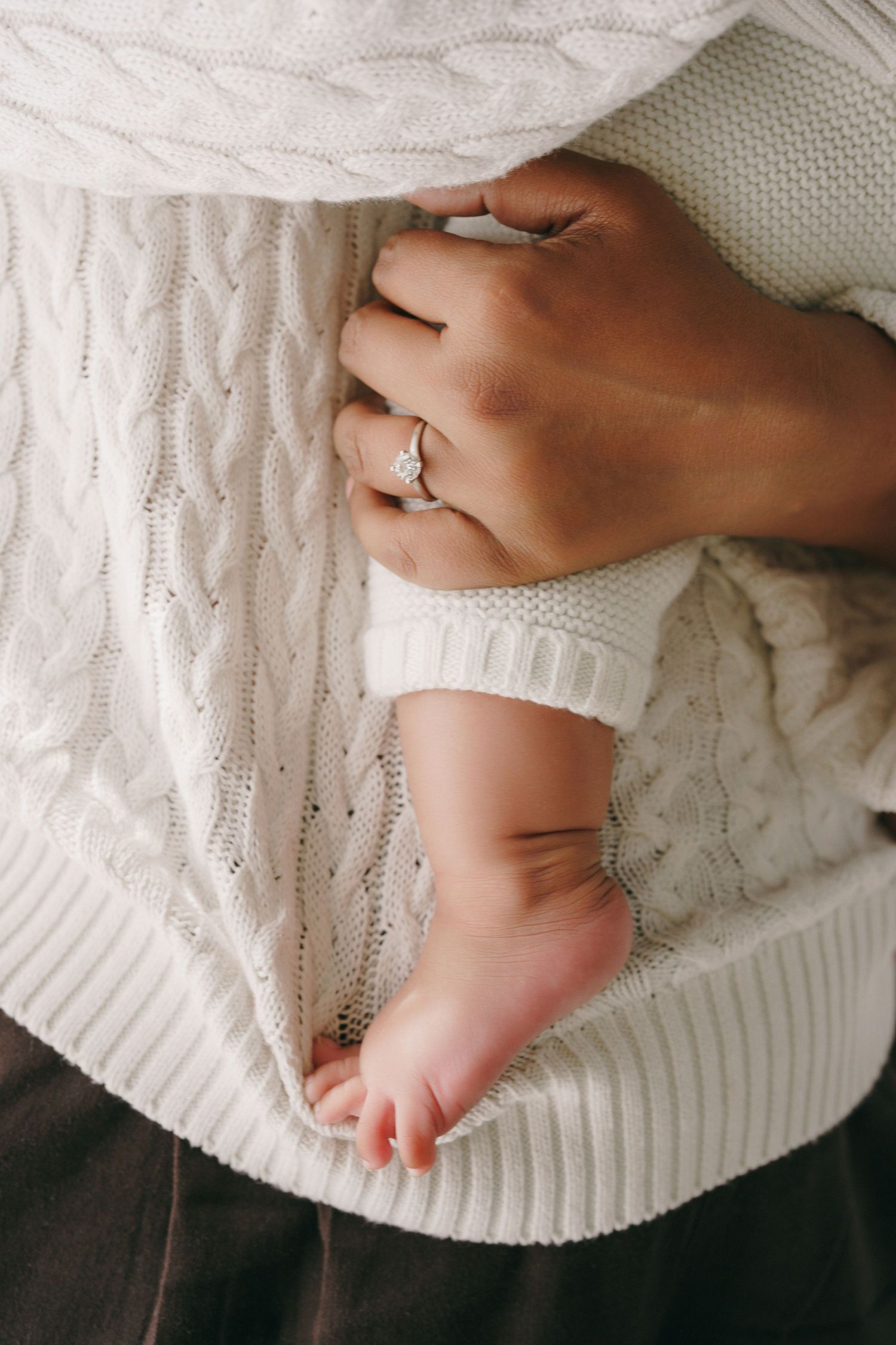 A mother wearing a diamond ring cradles her newborn baby's tiny feet in a cozy white cable-knit sweater.