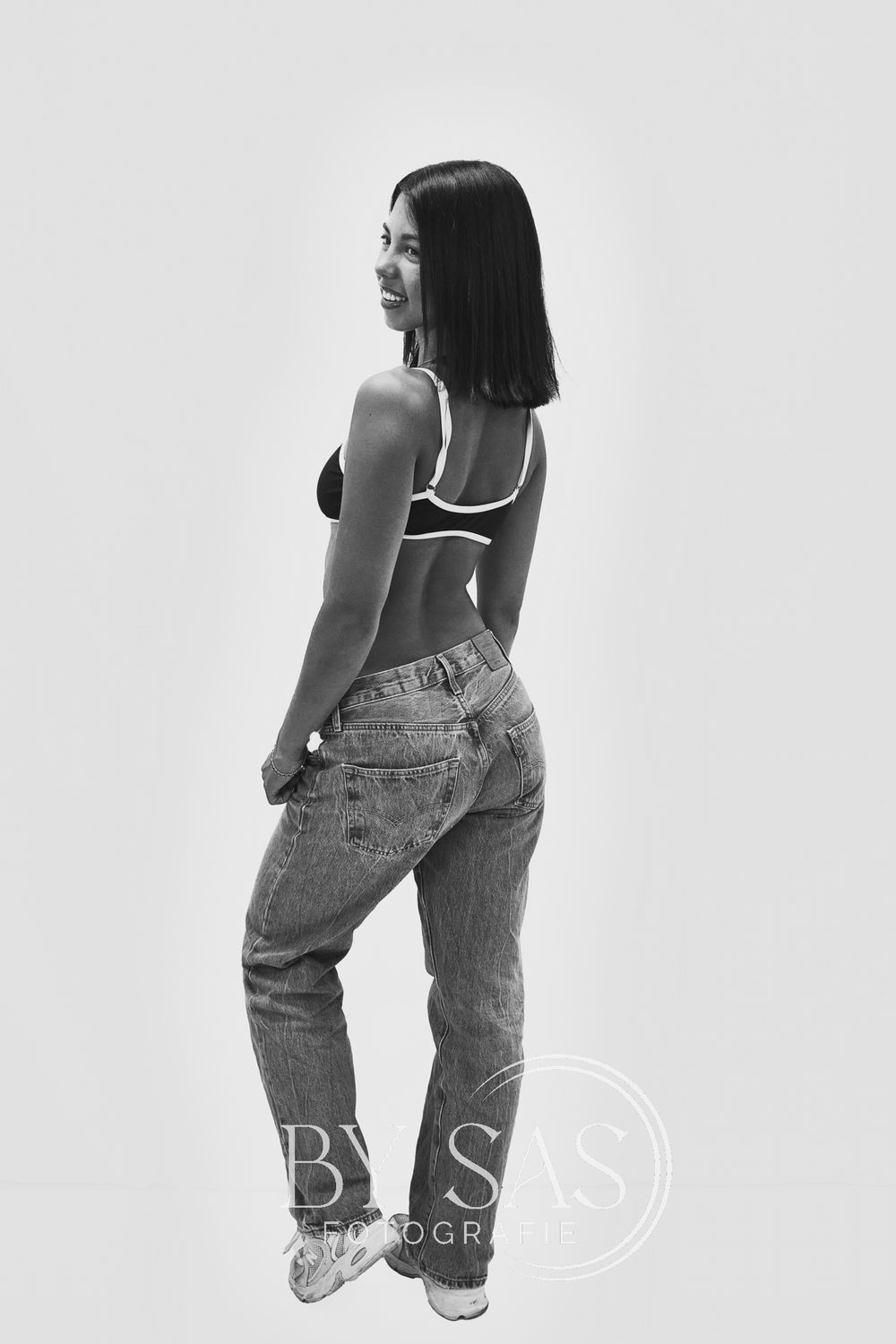 Black and white photo of a young woman in jeans and crop top posing from behind in a studio setting.