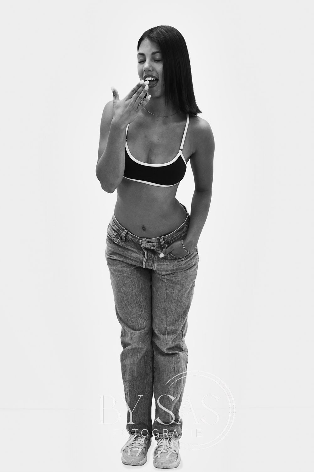 Black and white photo of a young woman in a crop top and jeans, posing with hand near her mouth.
