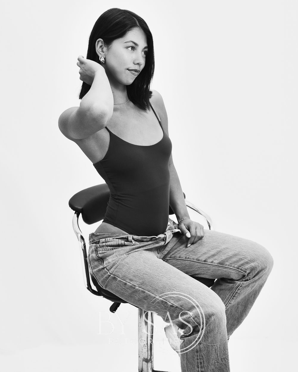 Black and white studio photo of a woman in a tank top and jeans sitting on a stool pointing forward.