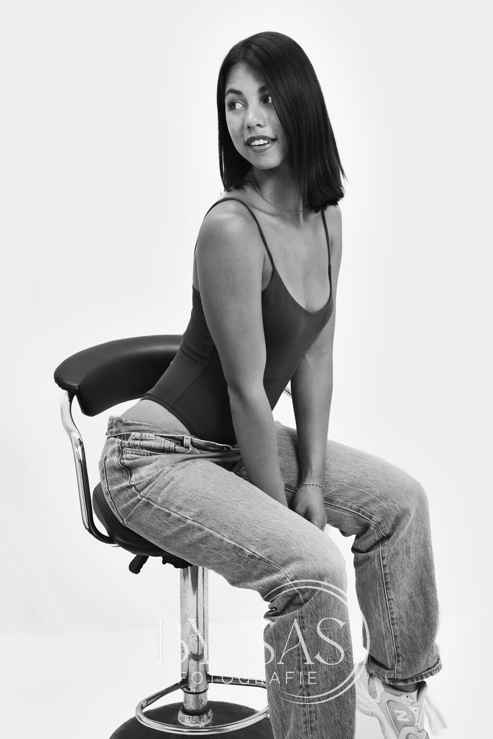 Black and white portrait of a woman in a bodysuit and jeans sitting on a bar stool in a studio.