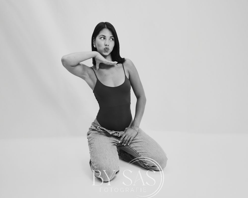Black and white photo of a young woman in a bodysuit and jeans posing on the floor.