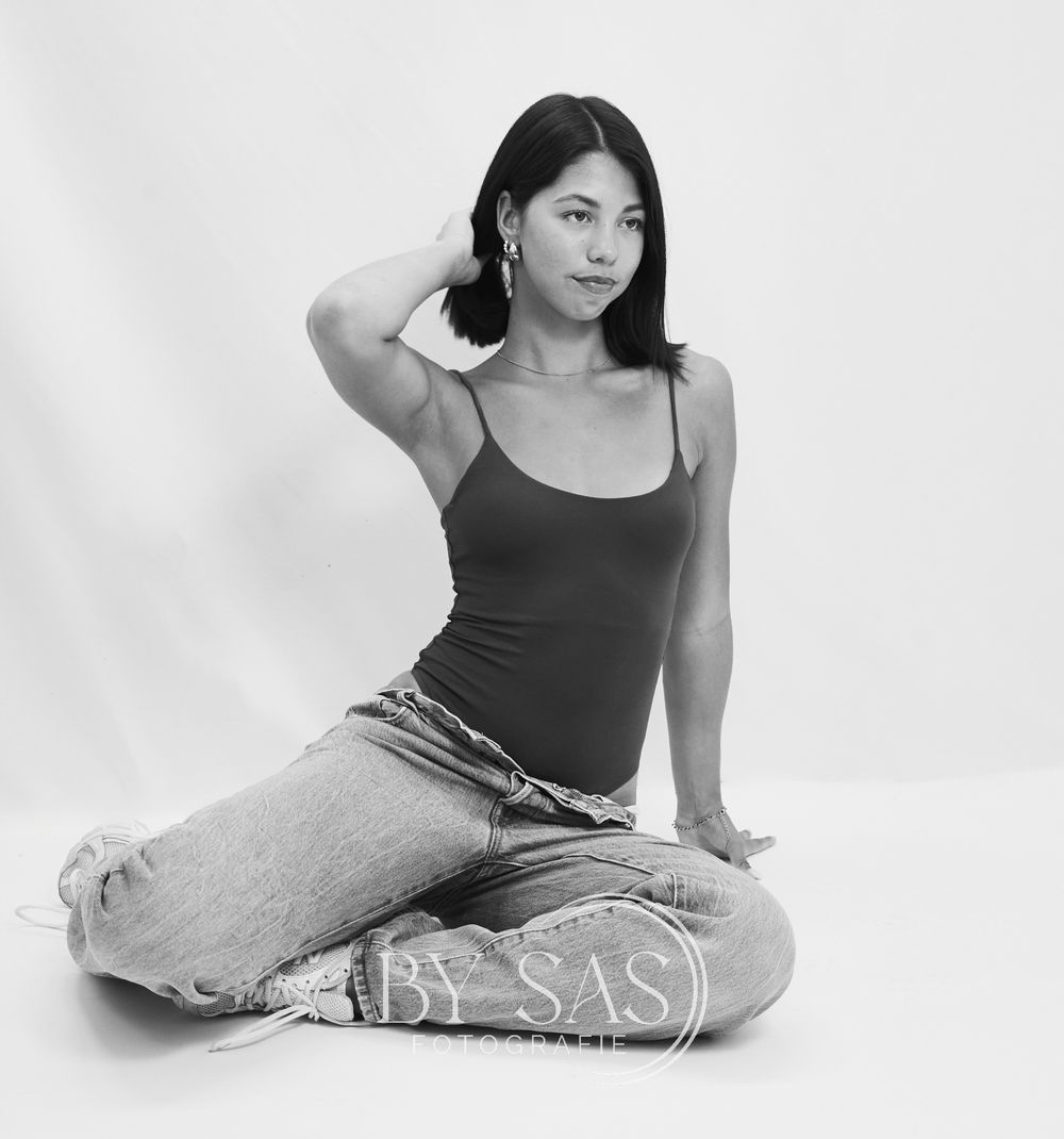 Black and white photo of a young woman sitting cross-legged in jeans and tank top, posing gracefully in a studio.