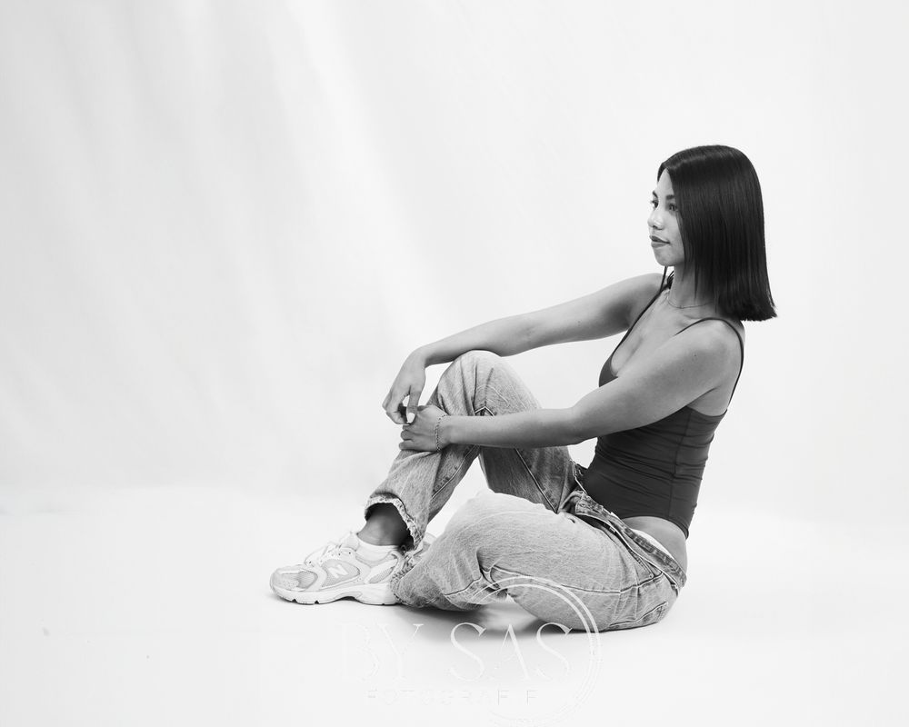 Black and white photo of a young woman sitting on floor in jeans and tank top, posing with knees up.