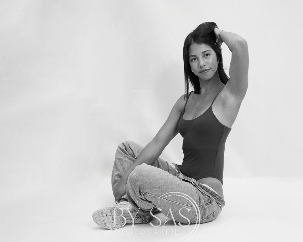 Black and white photo of a young woman sitting cross-legged on floor in bodysuit and jeans, posing with hand in hair.