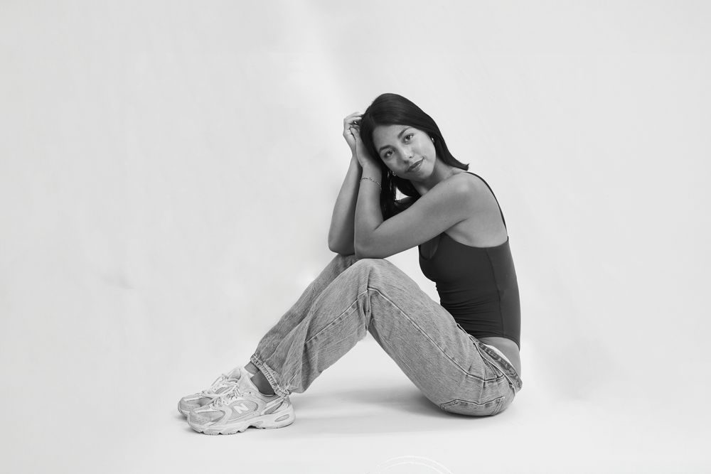 Black and white photo of a young woman sitting on floor in jeans and tank top, posing with knees up.