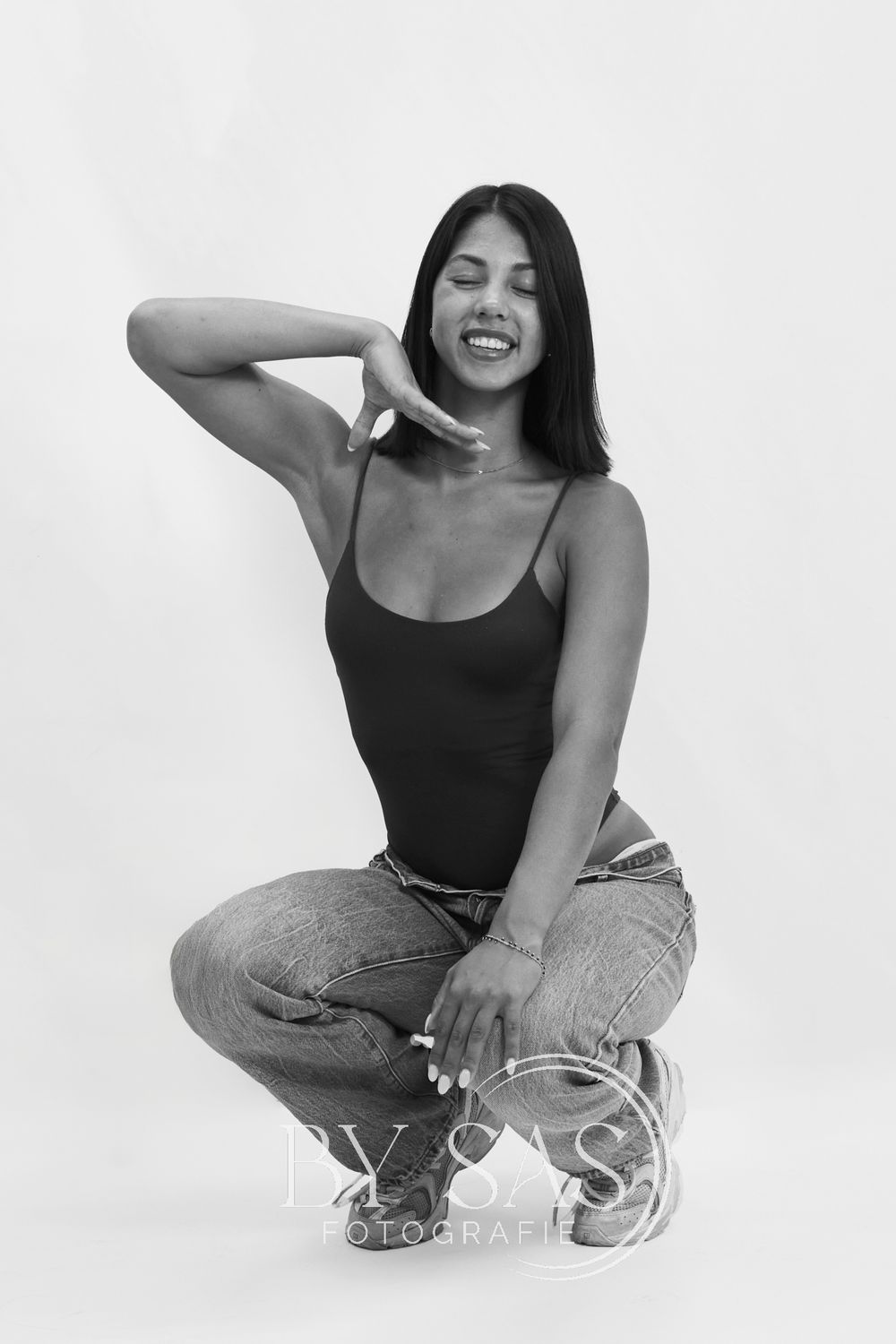 Black and white studio photo of a smiling woman in a bodysuit and jeans, posing in a squat position.