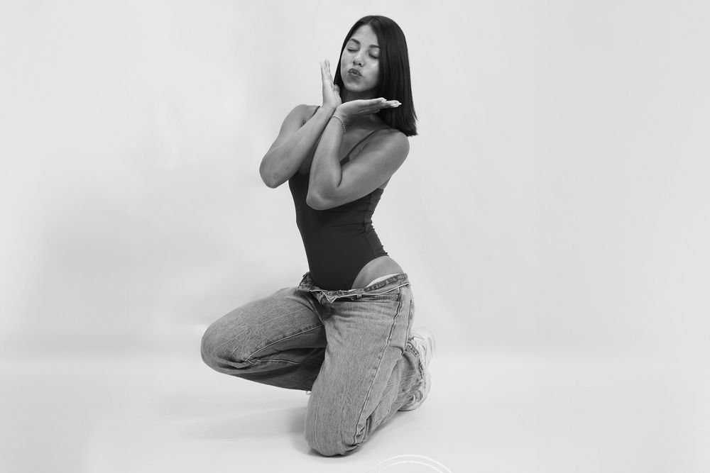 Black and white photo of a woman kneeling gracefully in jeans and bodysuit, posing with hands near face.