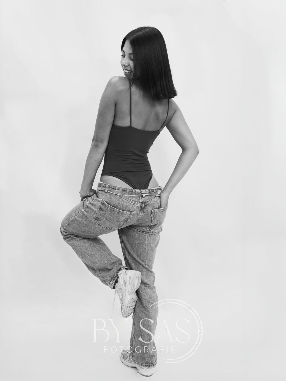 Black and white photo of a woman posing in jeans and a black tank top, looking over her shoulder.