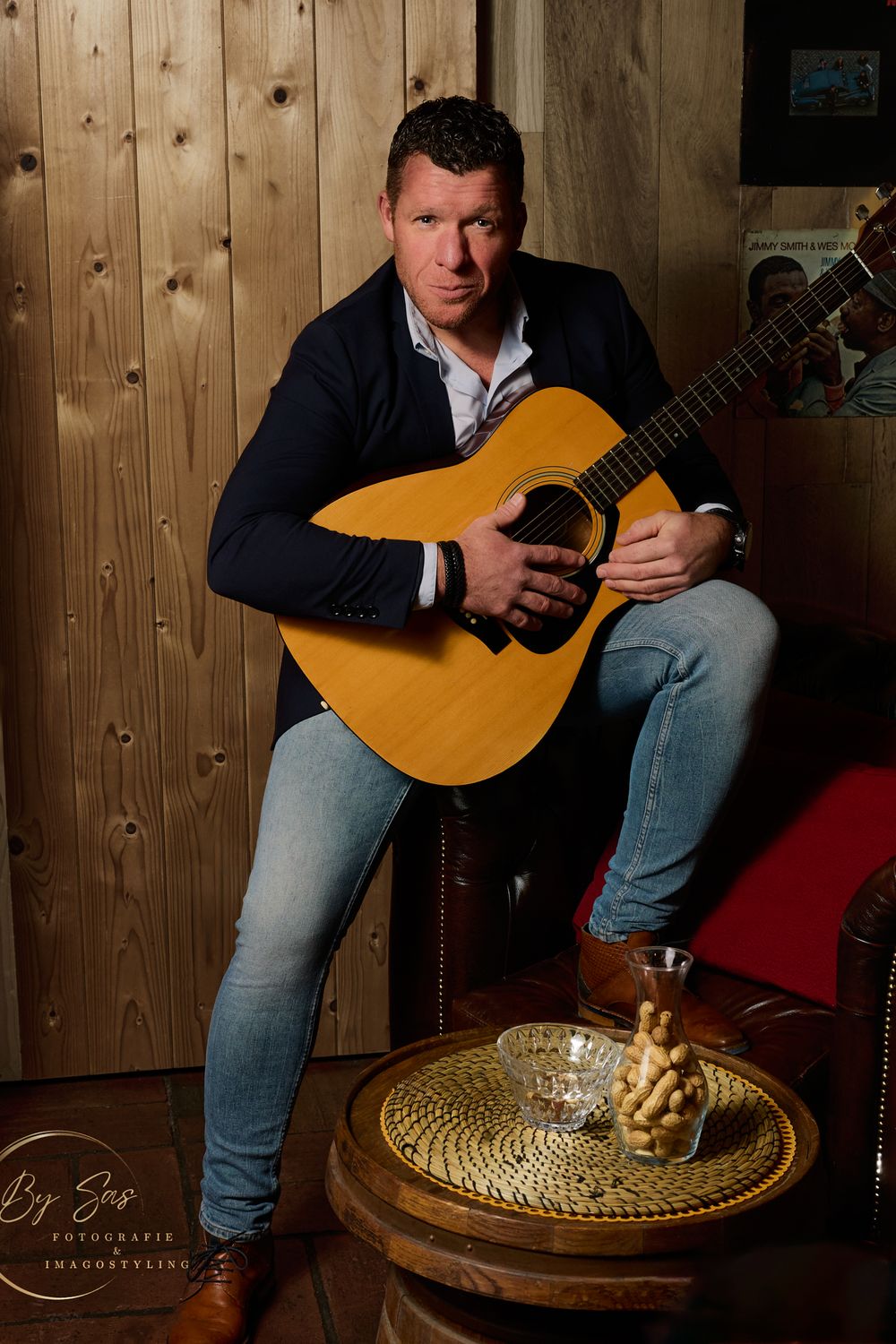 Man in navy cardigan playing acoustic guitar while seated in a rustic wooden interior setting.