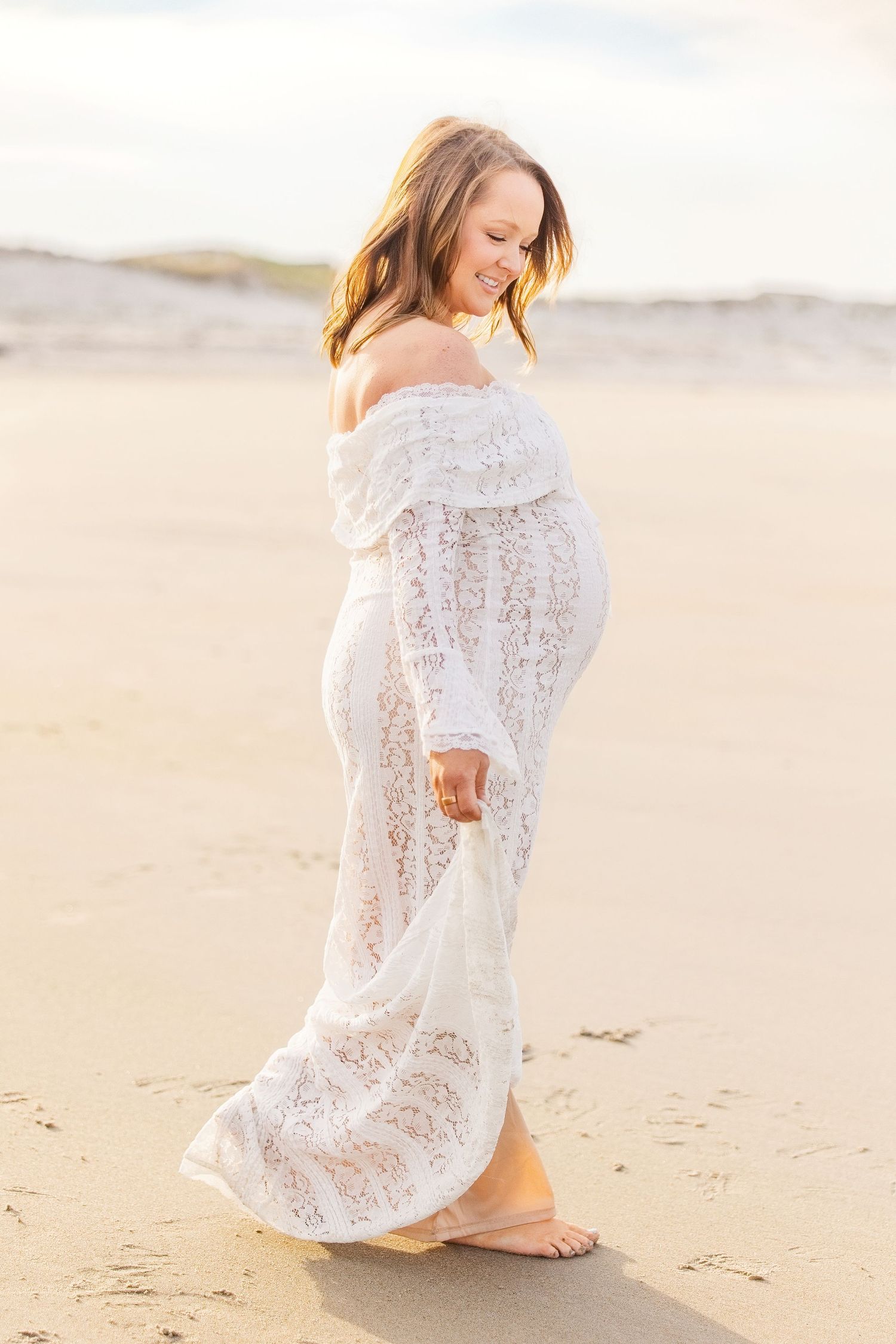 Pregnant woman in white lace off-shoulder gown walking barefoot on sandy beach at golden hour.