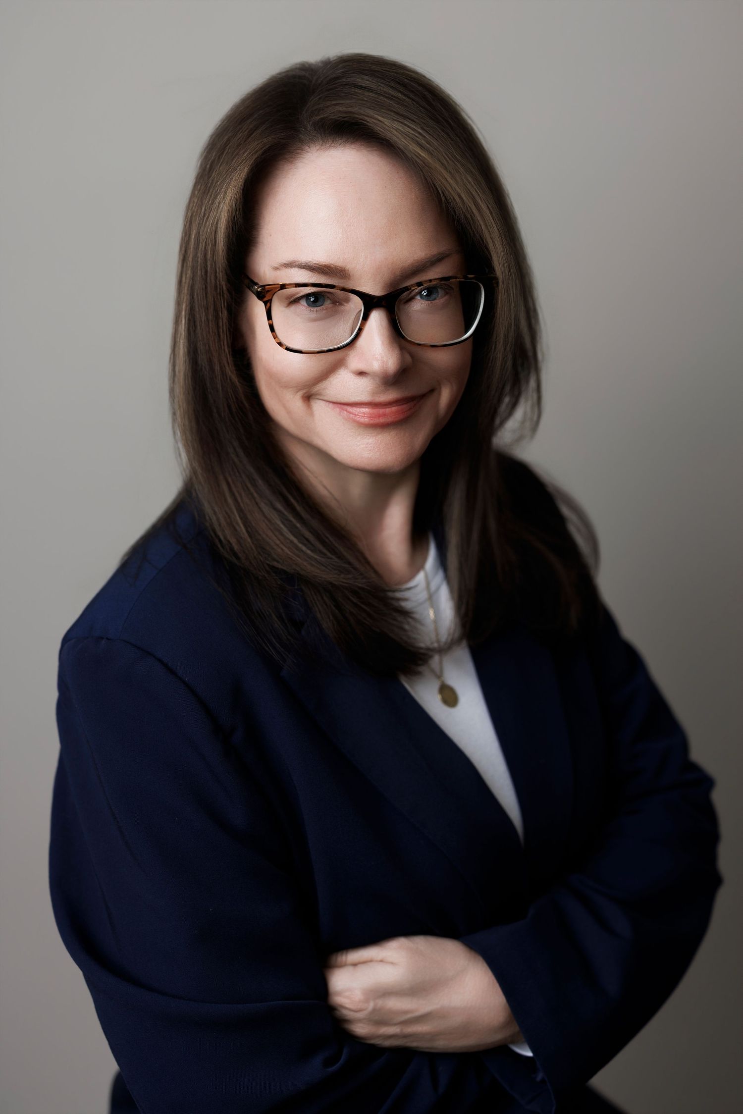 Professional woman with glasses and dark hair in navy blazer, arms crossed, smiling confidently against gray background.