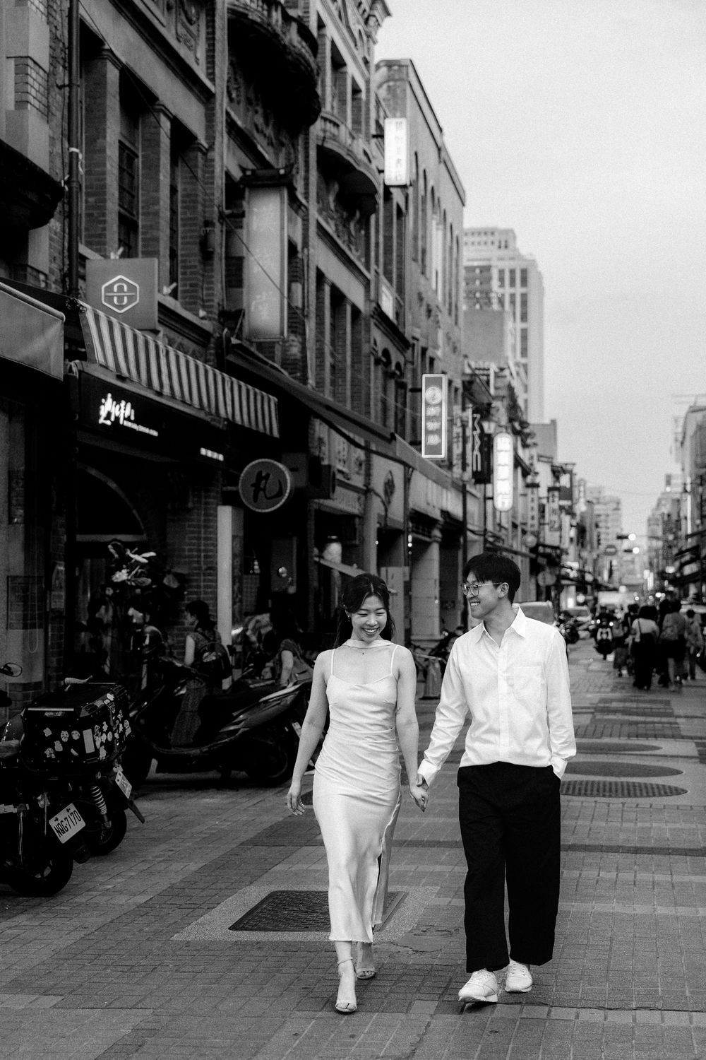 Black and white photo of a couple holding hands and walking along a busy urban street lined with shops and motorcycles.