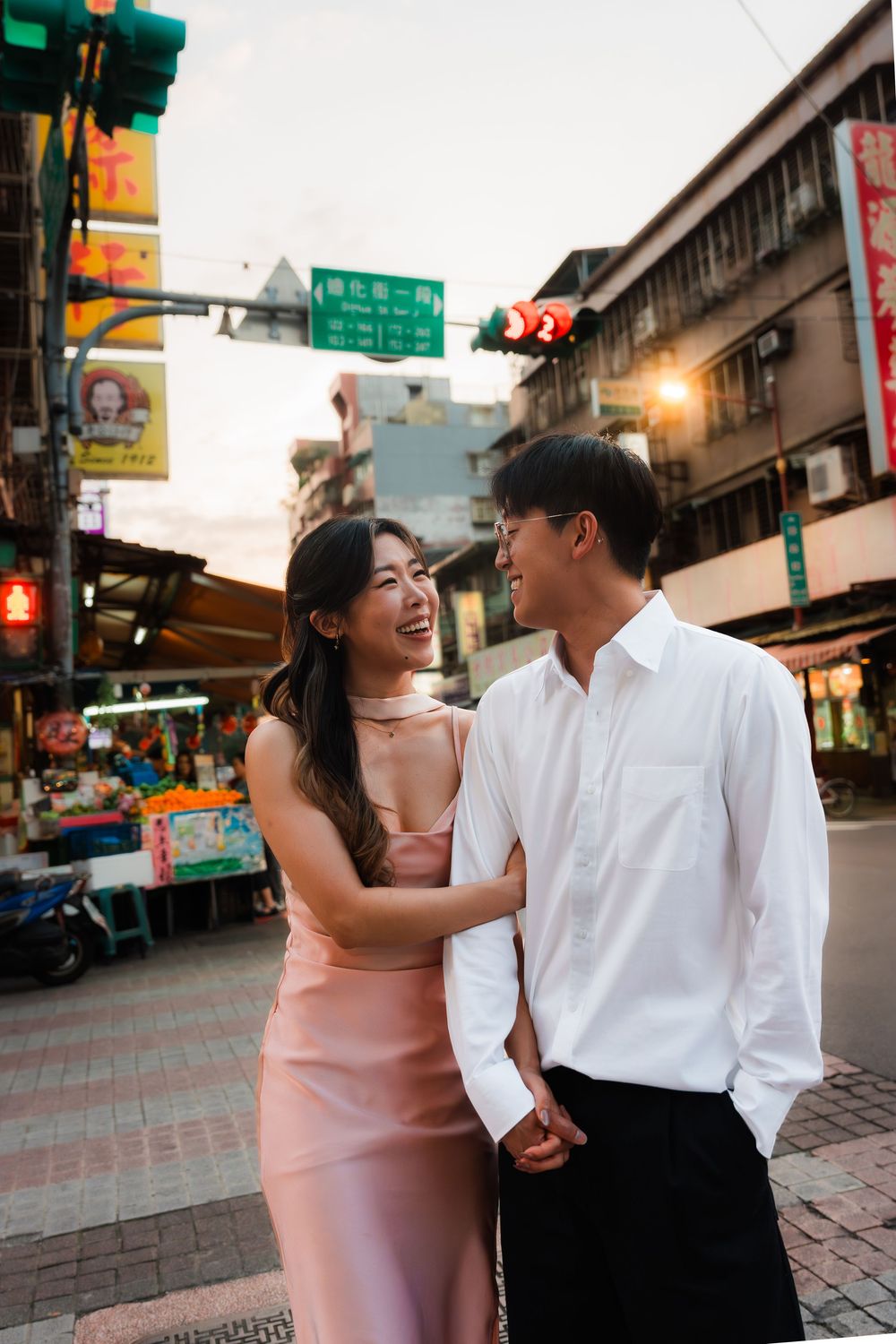Happy Asian couple holding hands and smiling at each other on a vibrant city street at dusk.