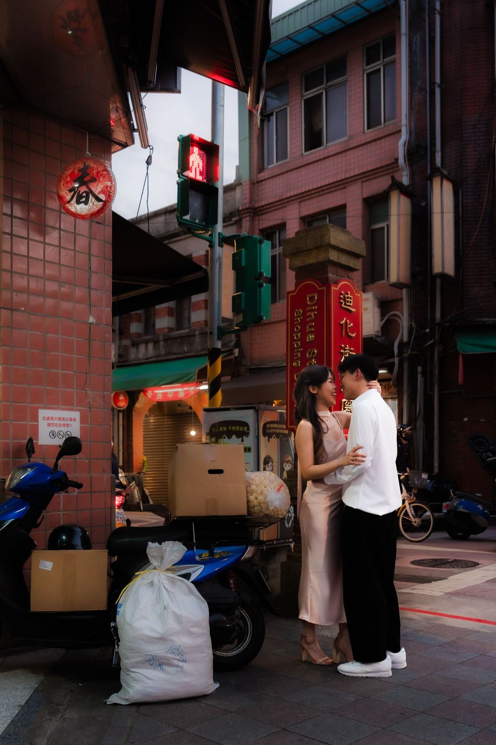 Couple embracing on a bustling Asian street corner with neon signs, scooters, and cardboard boxes nearby.