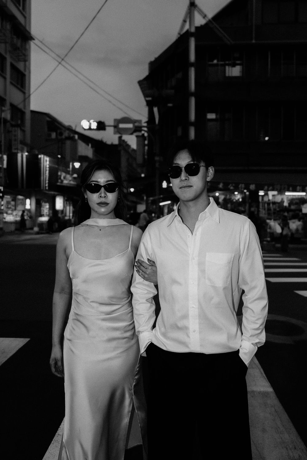 Black and white photo of a stylish couple in sunglasses posing on a city street at night.