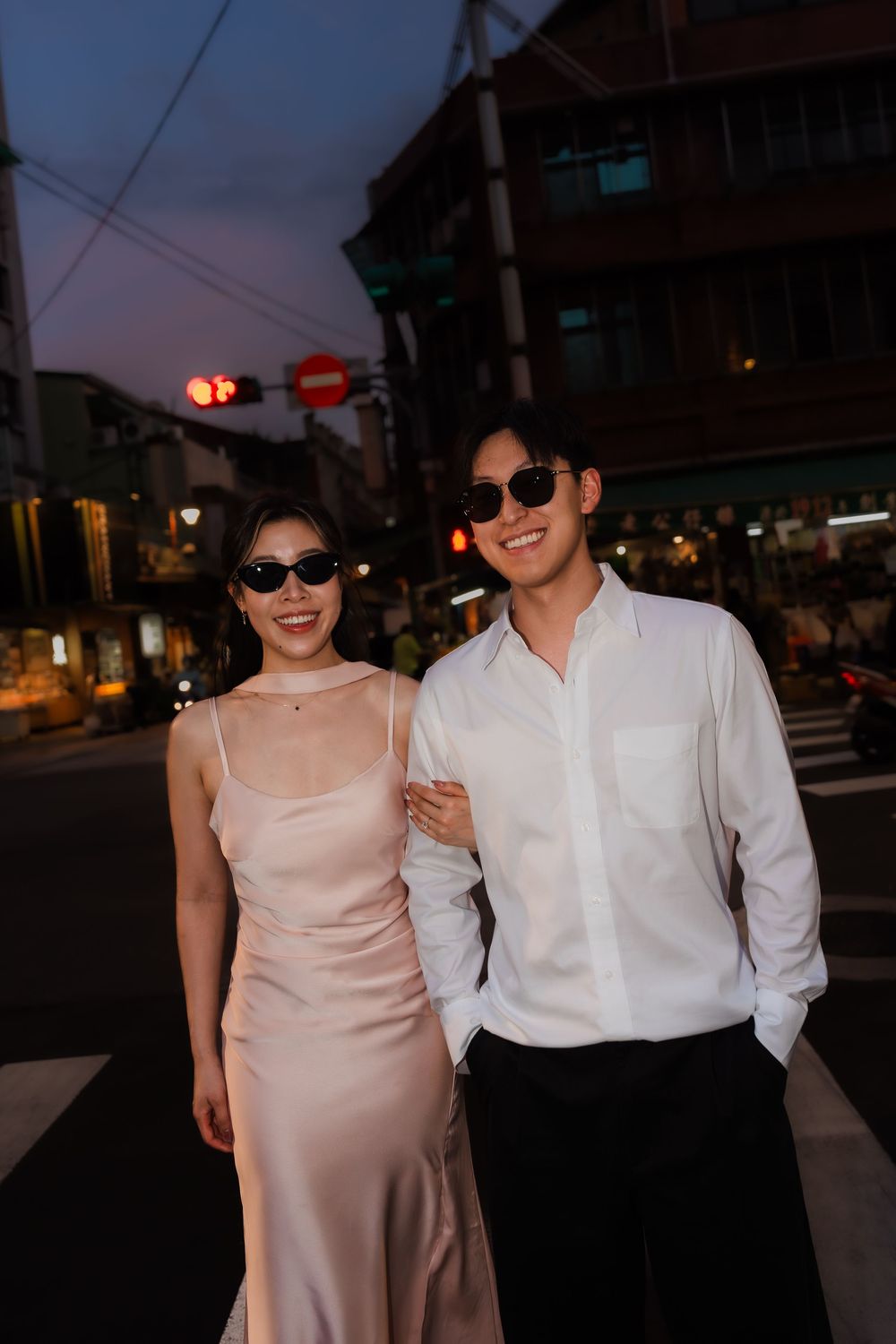 Stylish couple in sunglasses smiling on a city street at dusk, woman in satin dress, man in white shirt.