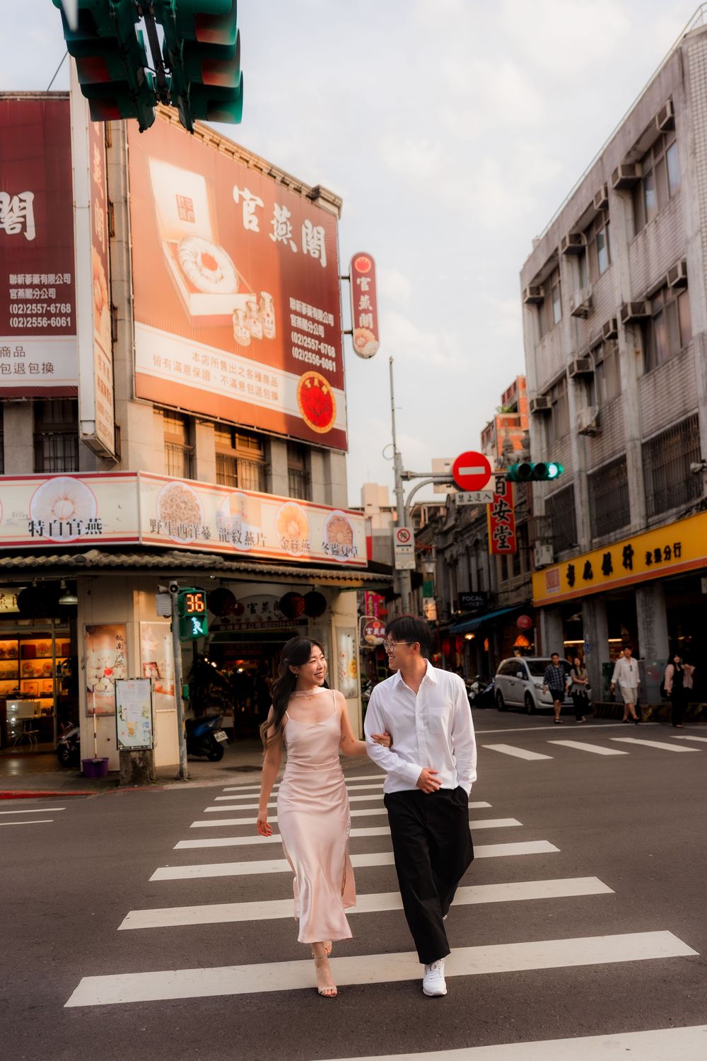 Couple walking hand in hand across a zebra crossing in a vibrant Taiwanese street with Chinese signage.