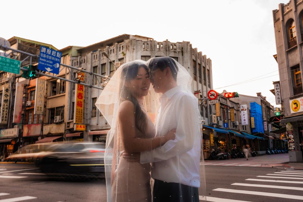 Bride and groom sharing a romantic kiss under a wedding veil at a busy Taiwanese street intersection.