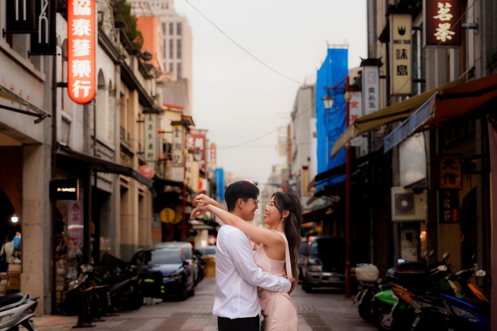 Couple dancing romantically in a bustling Asian street lined with shops and parked motorcycles at dusk.