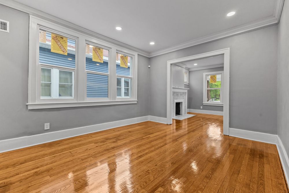 Bright living room with gleaming hardwood floors, gray walls, crown molding, and a view into an adjacent room with a fireplace. Detroit Mich