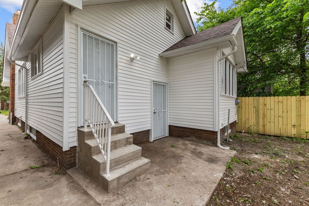 White siding home exterior with concrete steps, gray door, wooden fence, and lush green trees in background. Detroit Michigan Photo