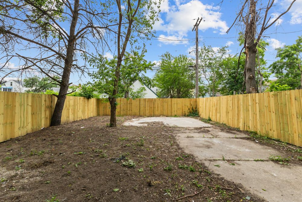 Backyard with new wooden privacy fence, bare dirt ground, concrete patio, and mature trees under a partly cloudy sky. Detroit Michigan Photo