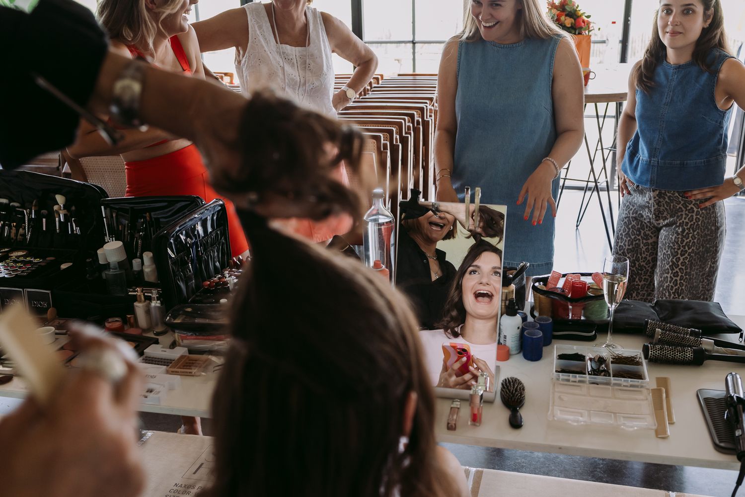 Women gathered around a makeup vanity table during a bridal getting-ready session, laughing and applying cosmetics.