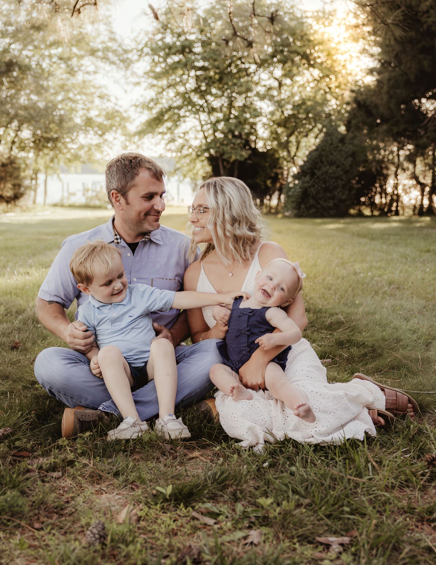 Happy family of four sitting on green grass in golden hour sunlight, parents smiling at each other with two young children.