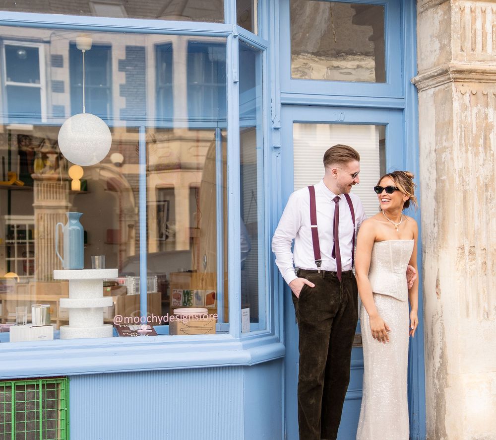 Bride and groom posing outside a blue-framed shop front, groom in suspenders smiling at bride in white dress.
