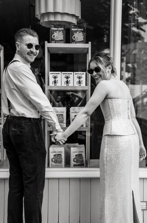 Black and white photo of a smiling couple holding hands in front of a coffee shop display, dressed in elegant wedding attire.